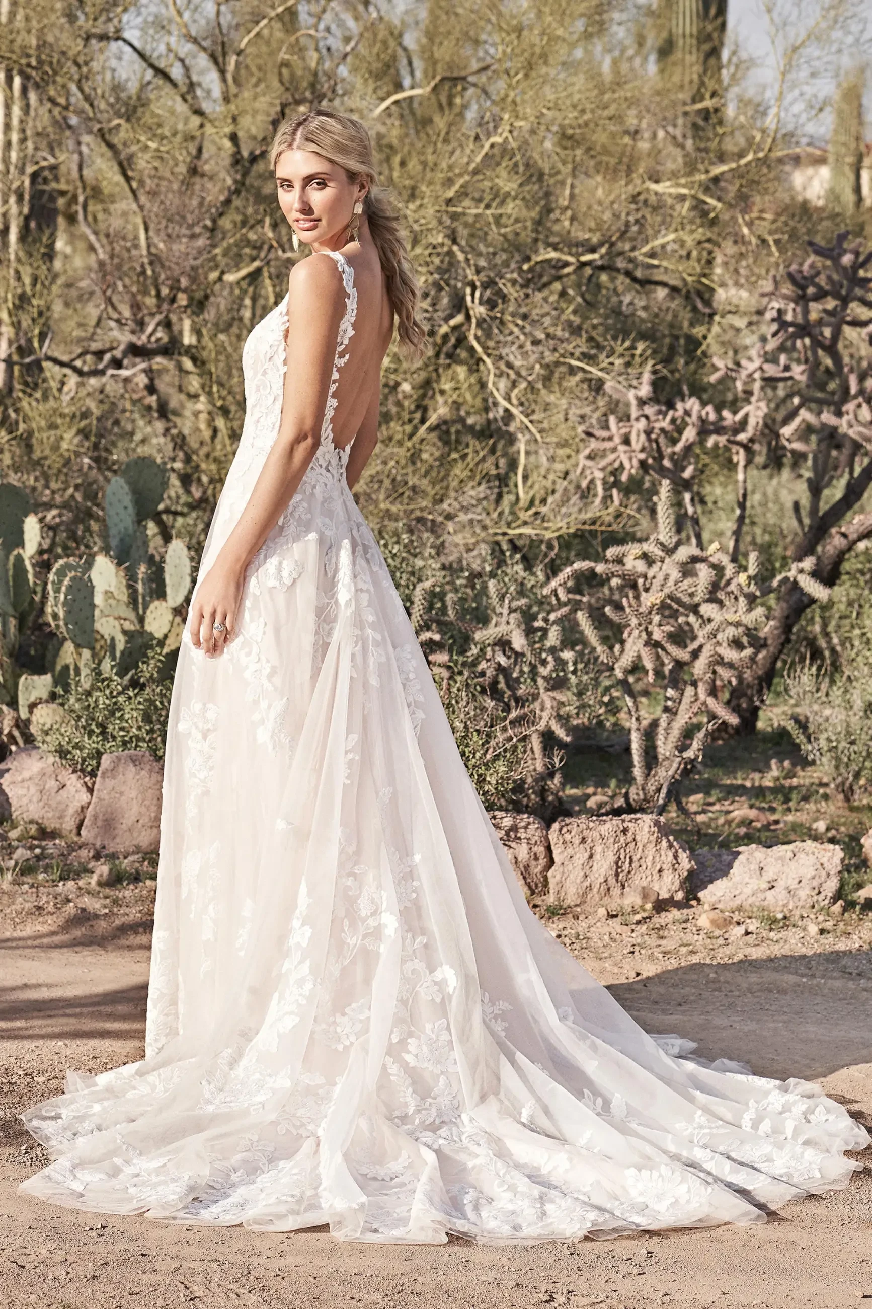 A woman in an elegant lace wedding dress stands outdoors, surrounded by desert plants and cacti. The scene conveys a sense of serenity and beauty.