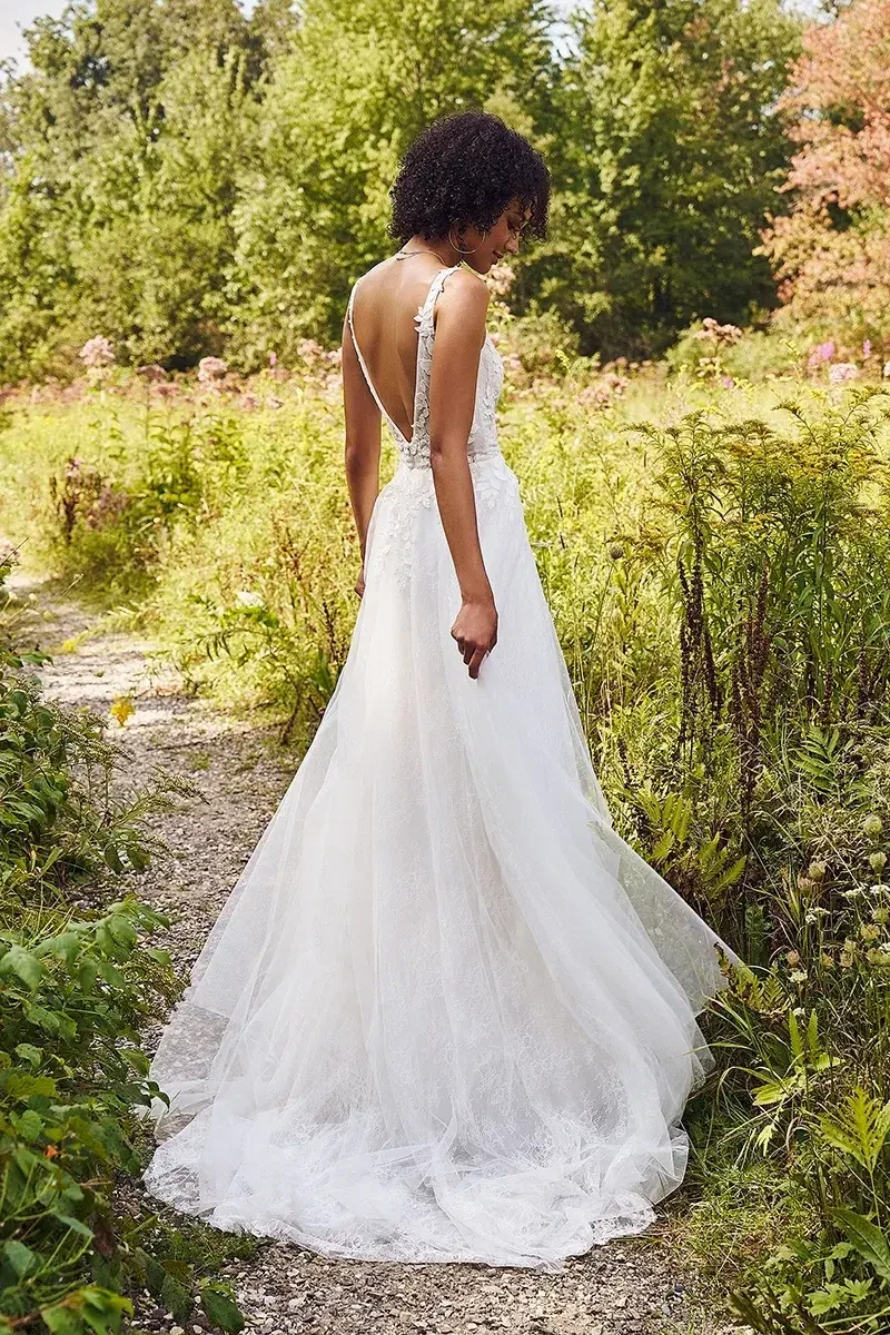 A woman in a white lace bridal gown with a slit walks on a path in a lush, green garden. She looks serene, surrounded by dense foliage and wildflowers.