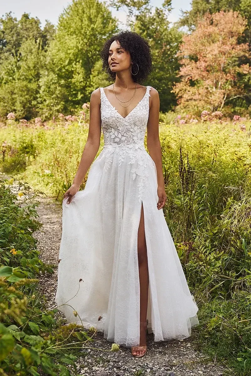 A woman in a white lace bridal gown with a slit walks on a path in a lush, green garden. She looks serene, surrounded by dense foliage and wildflowers.
