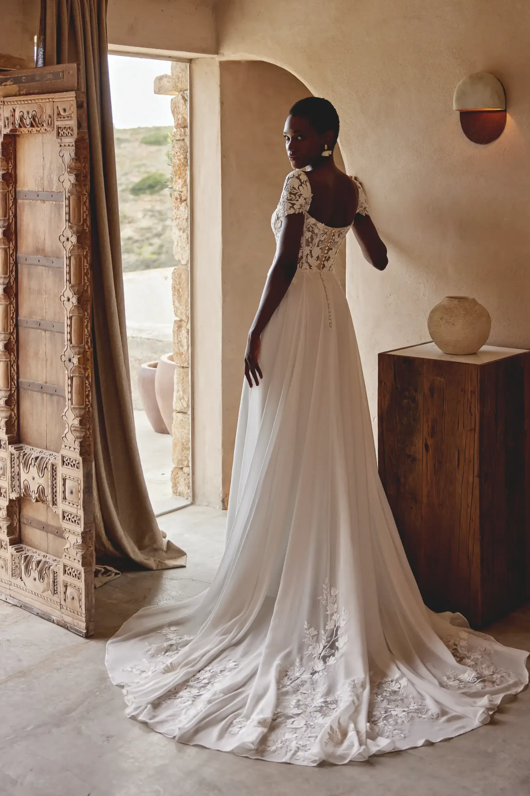 Elegant bride in a flowing white lace gown with cap sleeves stands in a minimalist room, exuding grace amidst rustic decor and warm tones.