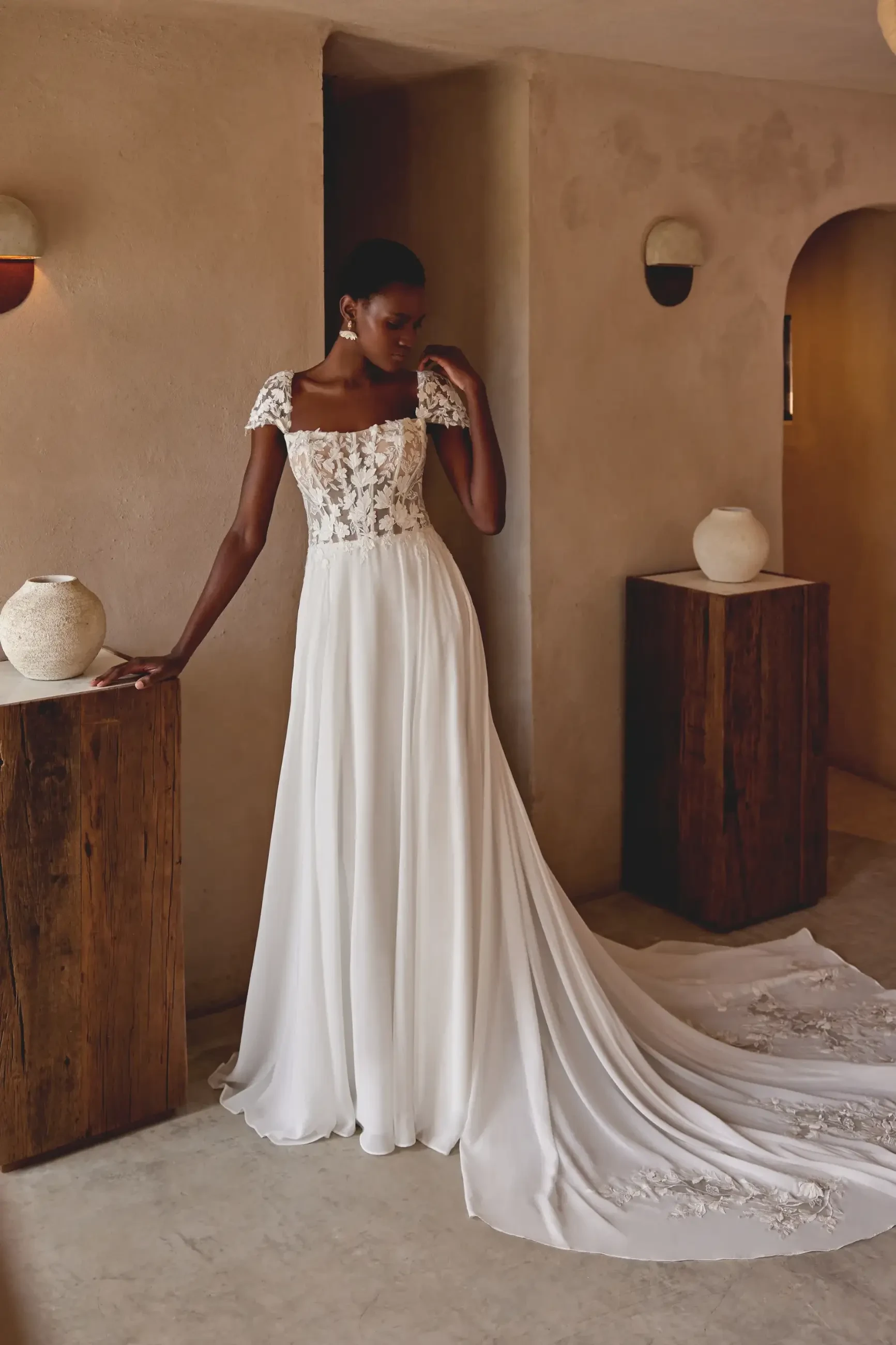 Elegant bride in a flowing white lace gown with cap sleeves stands in a minimalist room, exuding grace amidst rustic decor and warm tones.