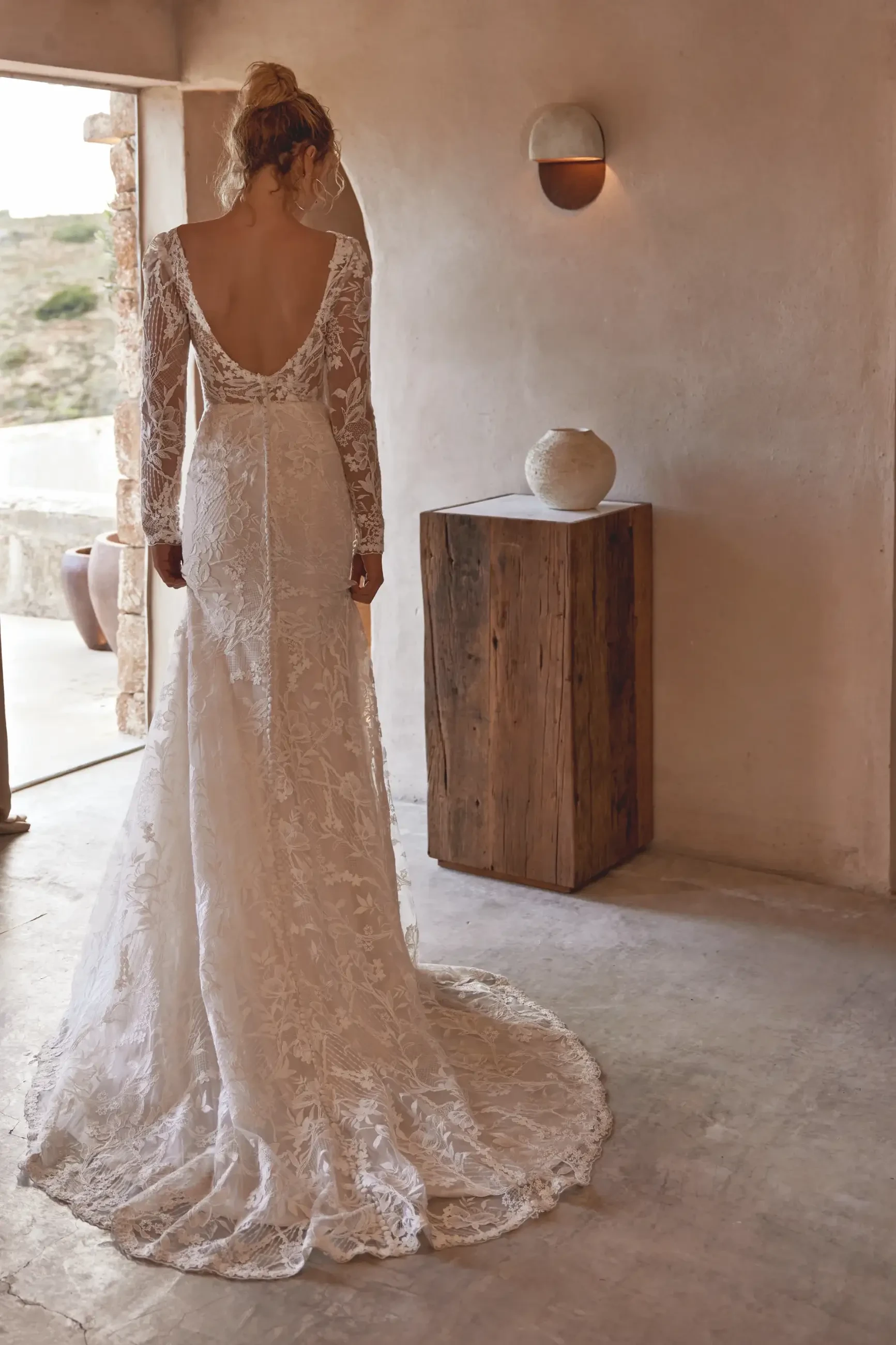 Bride in a lace wedding dress with long sleeves and a low back, stands in a rustic room with stone walls, wooden table, and soft, natural lighting.