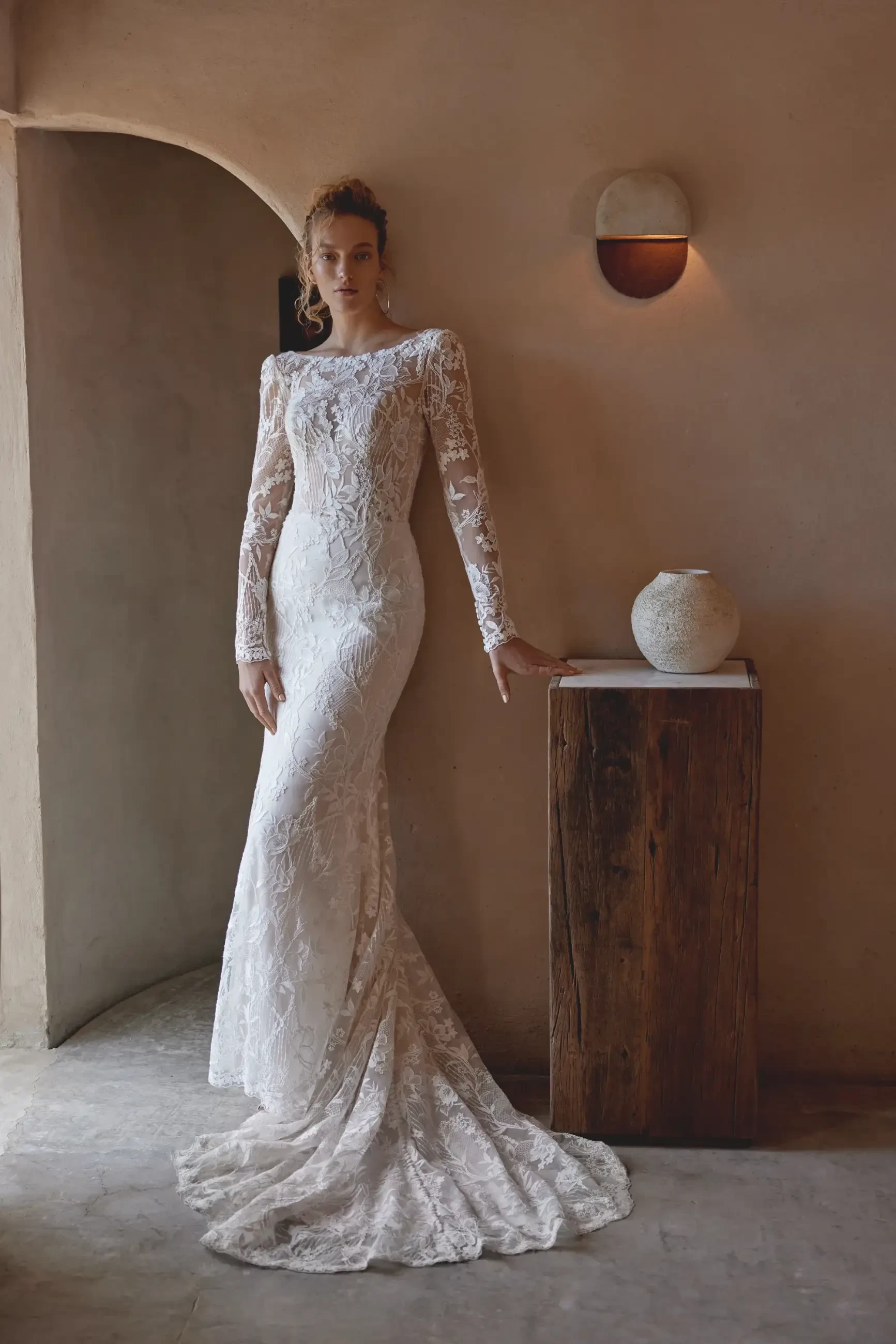 A woman in a long-sleeved, intricate lace wedding gown stands elegantly in a rustic room. She leans lightly on a wooden table with a white vase.