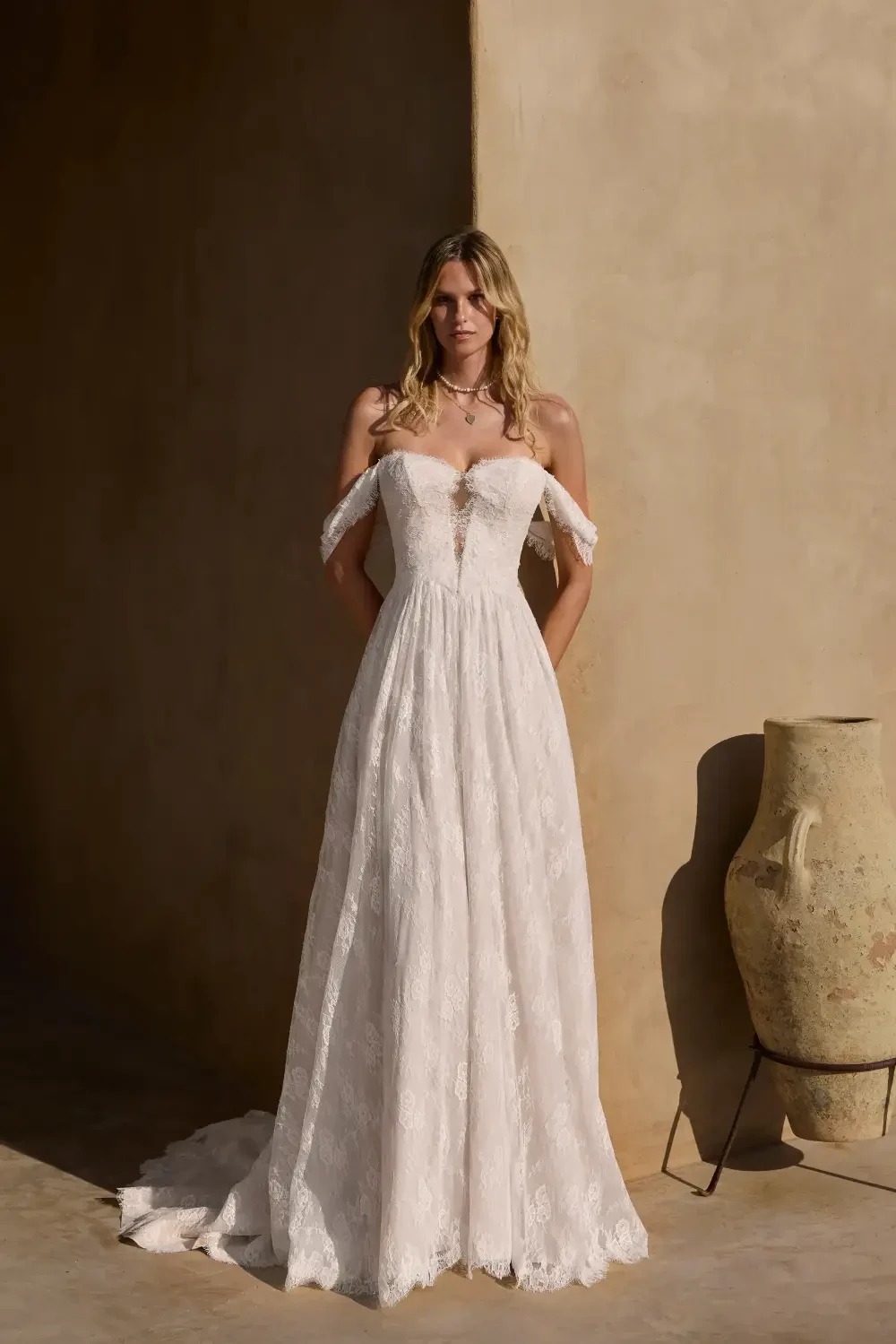 A woman wearing a strapless lace wedding dress, standing in front of a textured wall with a decorative vase nearby.