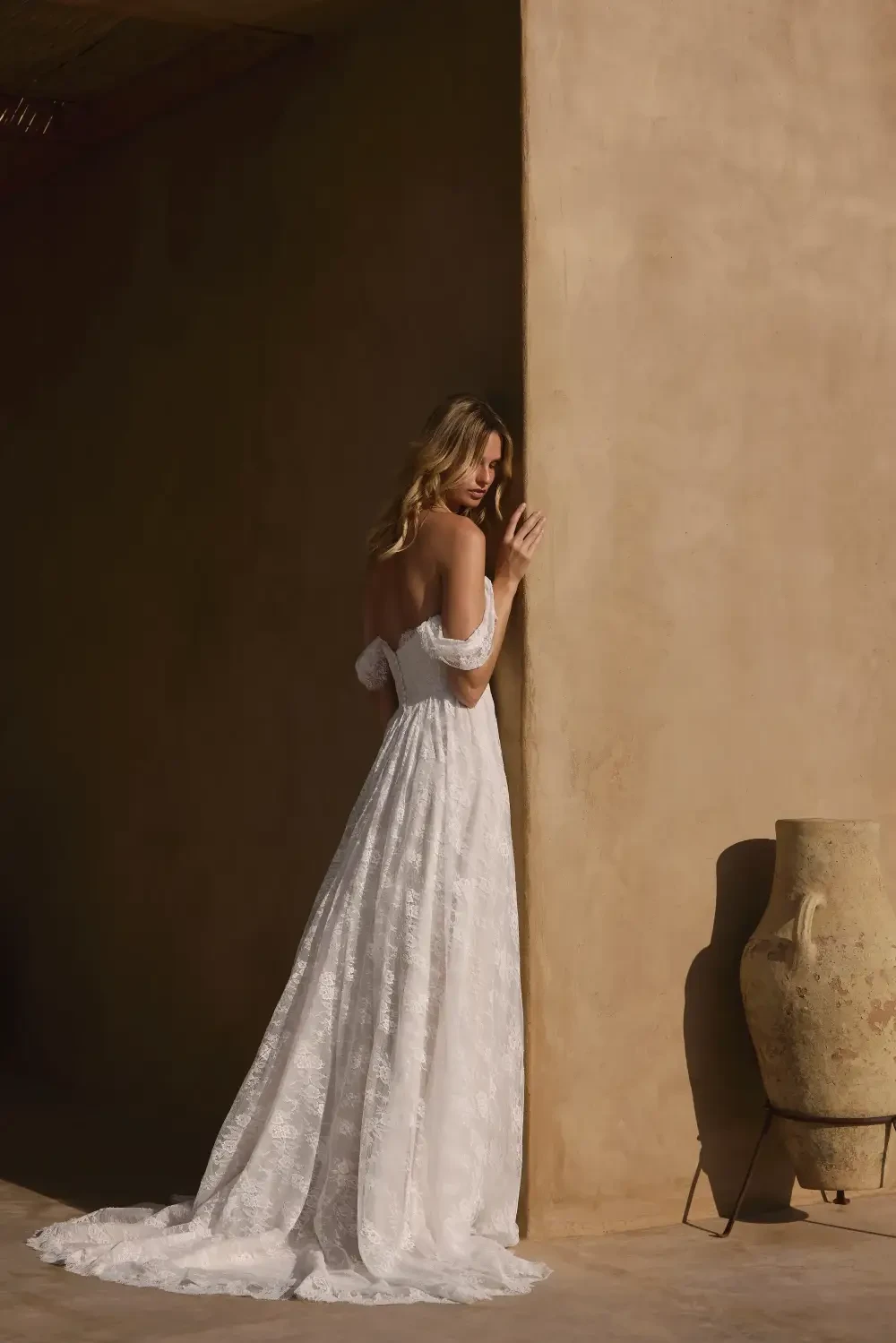 A woman wearing a strapless lace wedding dress, standing in front of a textured wall with a decorative vase nearby.