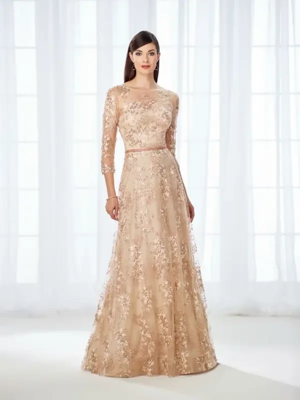 A model stands in a light-filled room wearing an elegant, floor-length gown adorned with delicate floral embroidery. The dress has three-quarter length sleeves and a fitted bodice, complementing her graceful posture.
