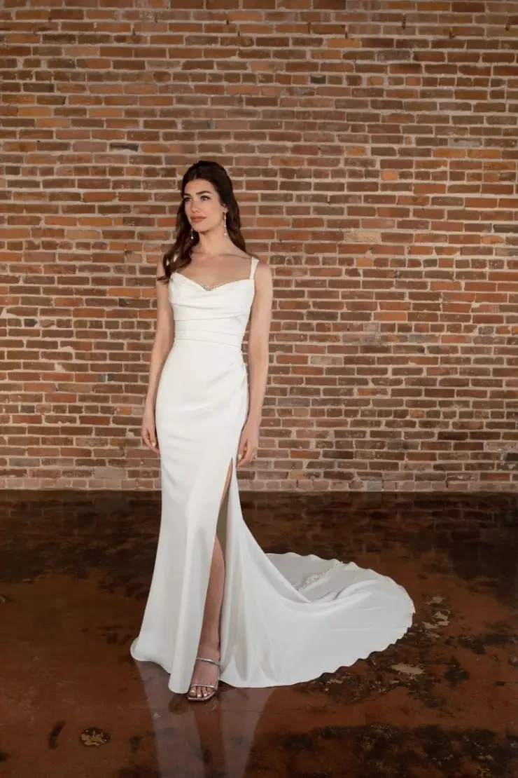 A woman stands elegantly in a white, sleeveless gown with a long train and side slit. She is posed against a textured brick wall, exuding a sophisticated and serene vibe.