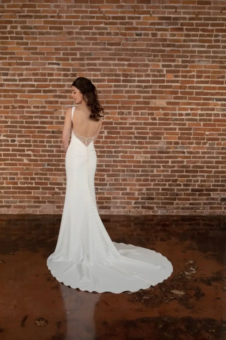 A woman stands elegantly in a white, sleeveless gown with a long train and side slit. She is posed against a textured brick wall, exuding a sophisticated and serene vibe.