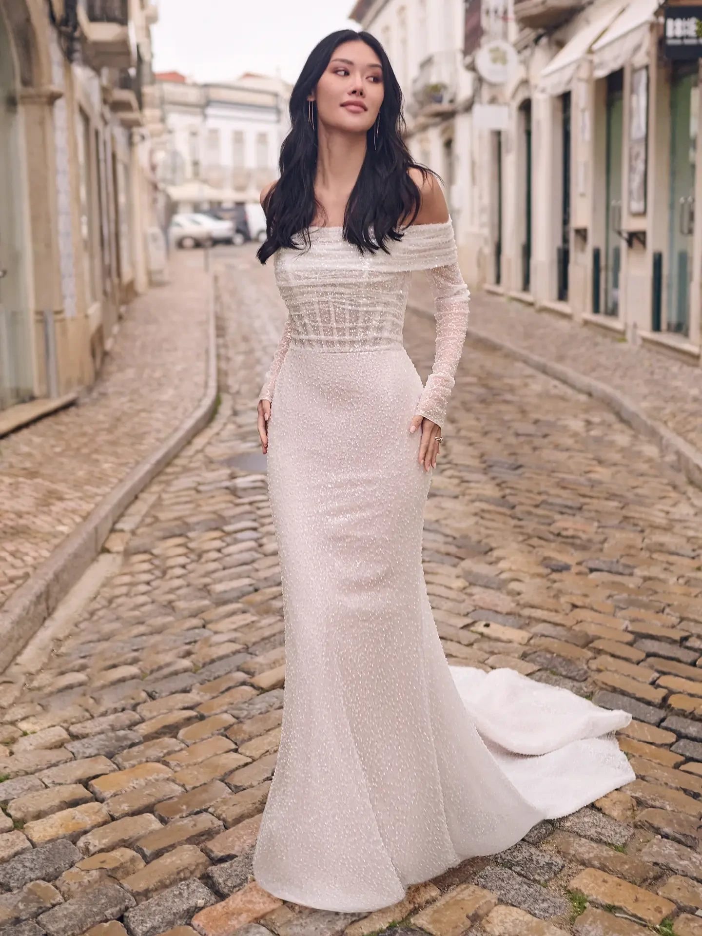 Woman in an elegant, off-shoulder white gown stands on a cobblestone street lined with rustic buildings, conveying a serene, timeless vibe.