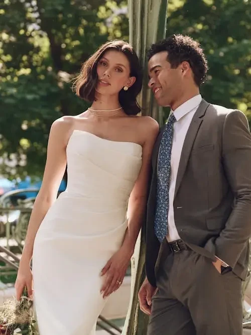 A bride in a strapless white gown leans against a tree, smiling softly. Beside her, a groom in a gray suit stands with a relaxed, joyful expression. Trees and soft lighting create a serene outdoor setting.