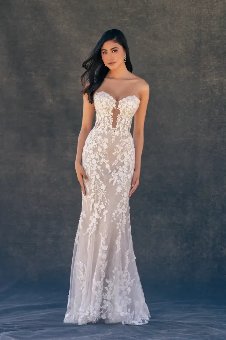 A woman stands elegantly in a strapless, lace wedding gown with floral patterns and a sheer bodice against a dark, textured background, exuding sophistication.
