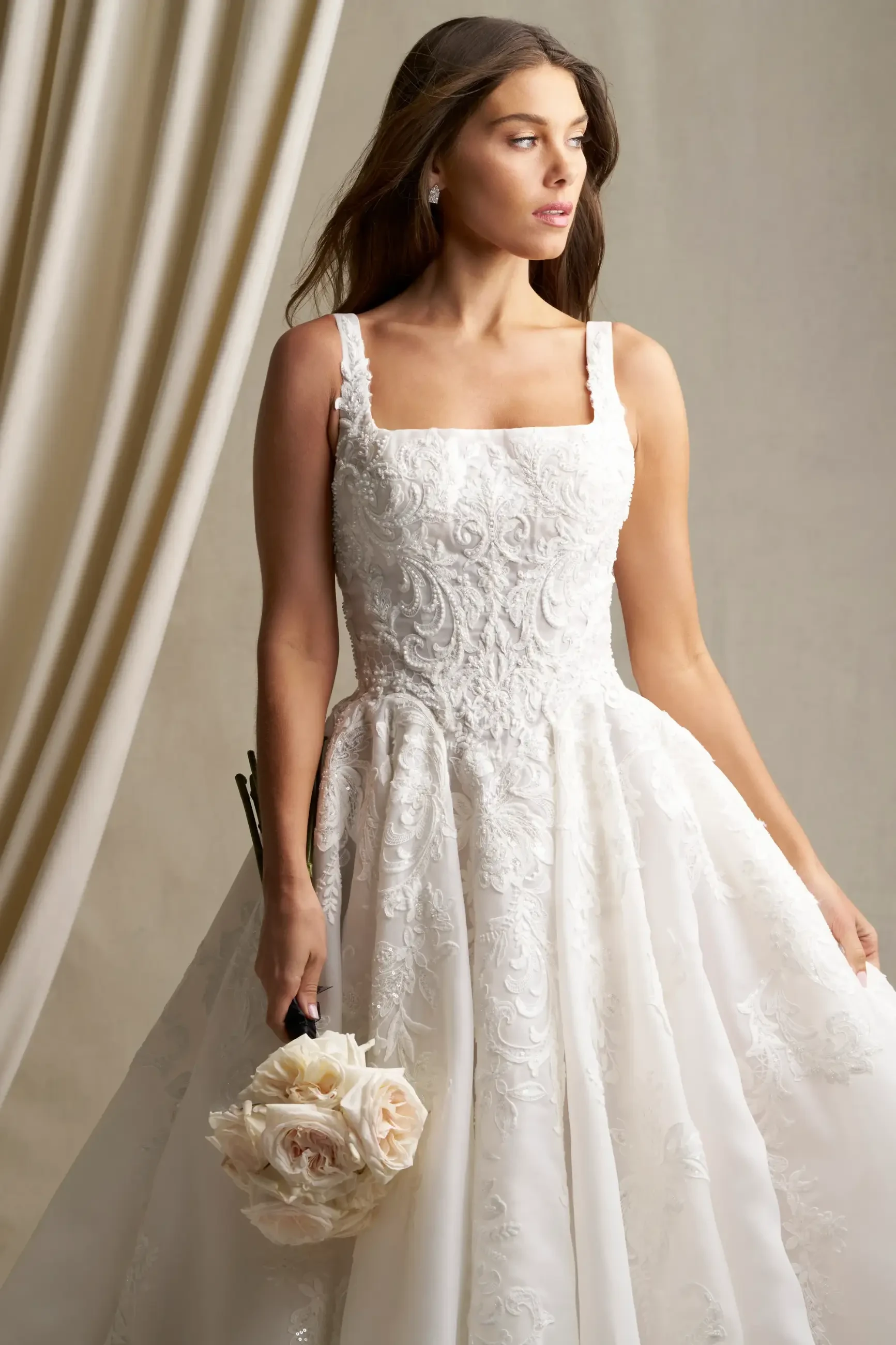 A woman in a white wedding dress with a square neckline holds a bouquet of flowers and poses against a neutral background.