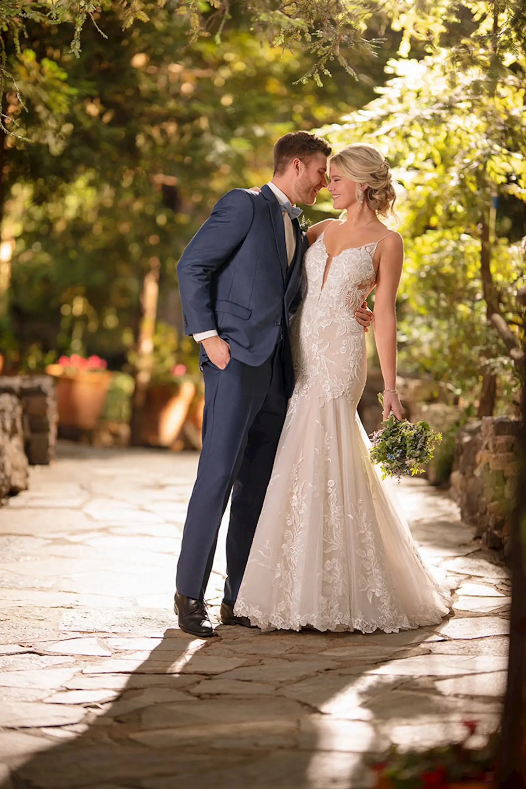 A bride in a lace gown and a groom in a blue suit stand closely on a sunlit stone path, surrounded by lush greenery, conveying romance and joy.