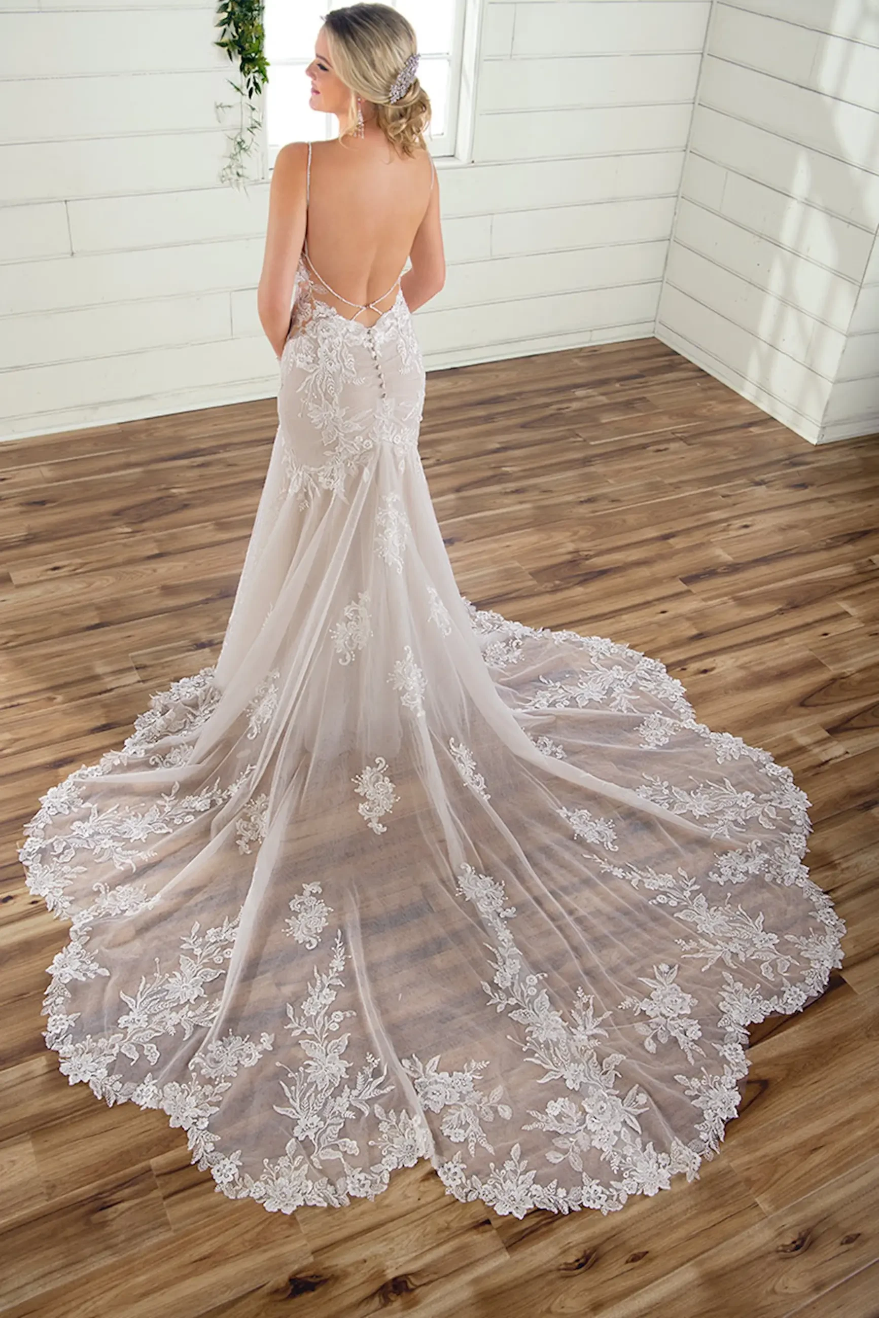 Bride in backless lace gown with intricate floral patterns stands on polished wooden floor, exuding elegance in sunlit, white-paneled room.