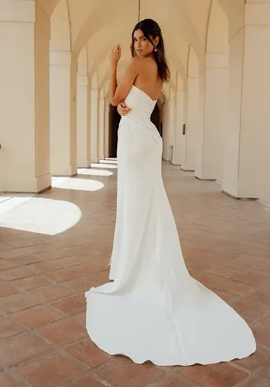 A woman in a strapless white gown stands elegantly in a sunlit, arched hallway. Her expression is serene, with soft lighting creating a romantic ambiance.