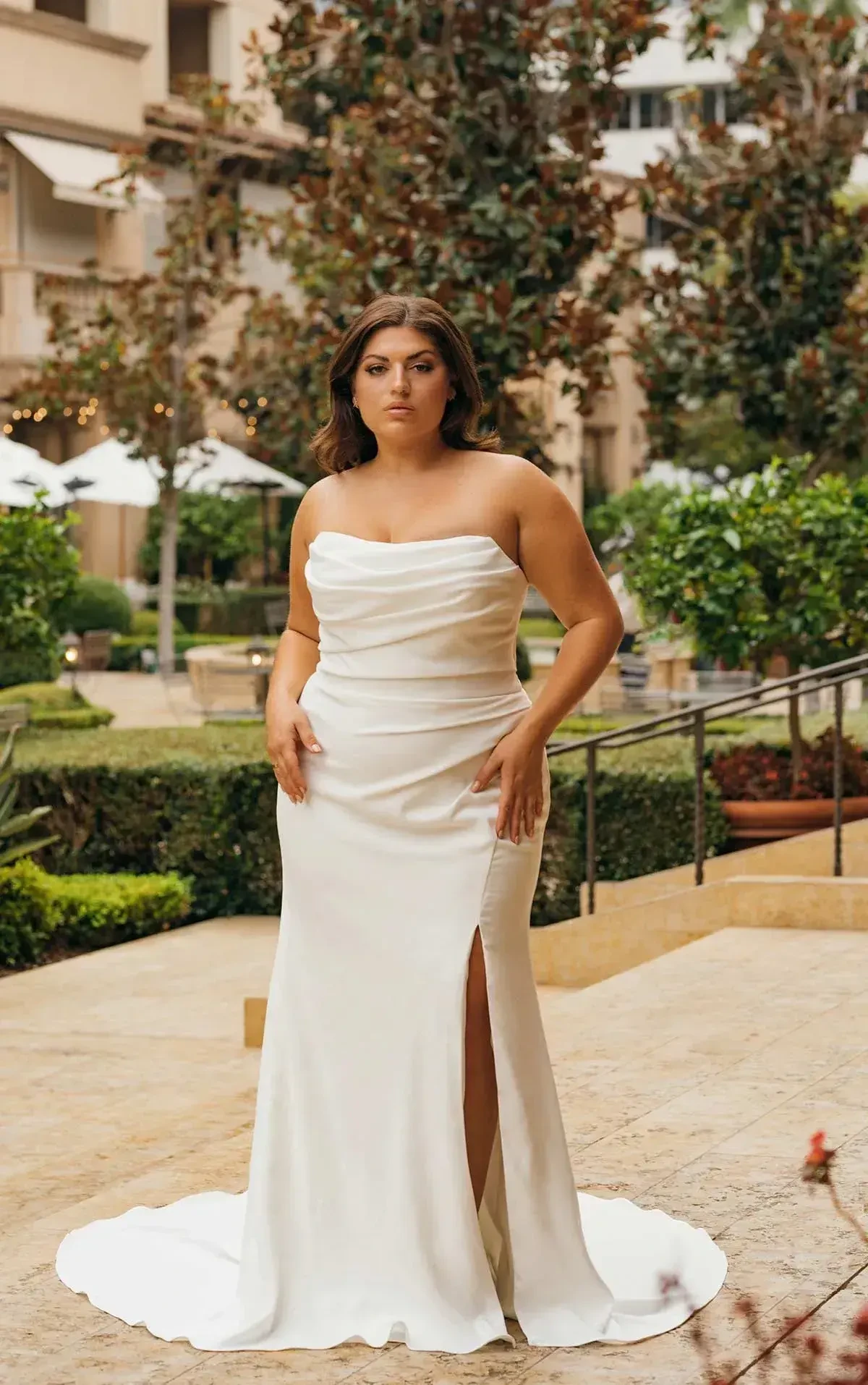 Woman in a strapless white gown stands confidently in a garden courtyard, surrounded by lush green plants and soft outdoor lighting, exuding elegance.