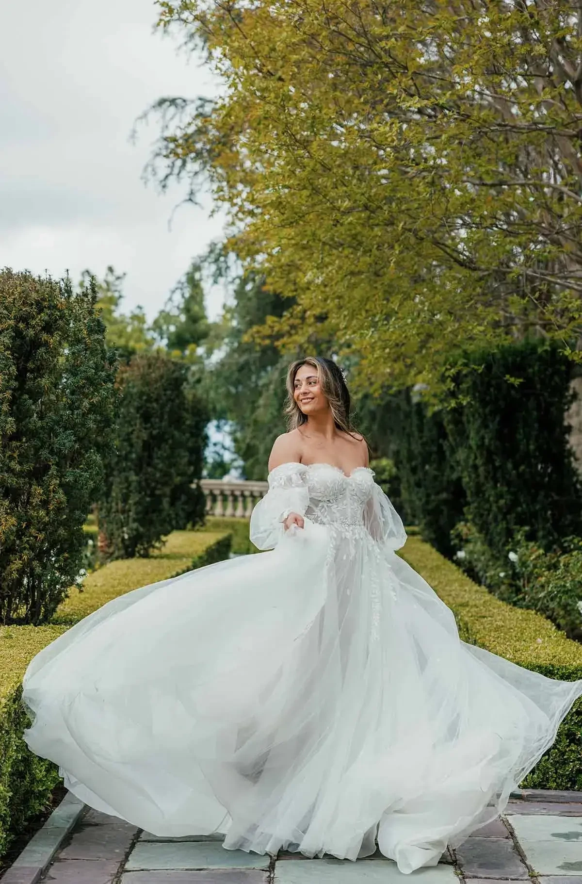A woman twirls in a flowing off-the-shoulder wedding dress in a lush garden. Her expression is joyful, surrounded by manicured hedges and trees.