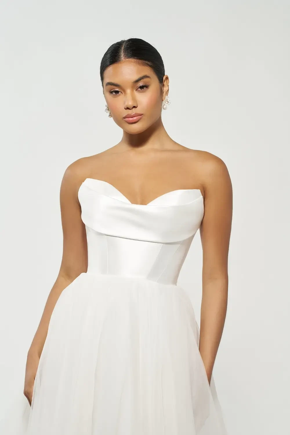 A woman is wearing a strapless white dress with a fitted bodice and a full tulle skirt, posing against a plain background.