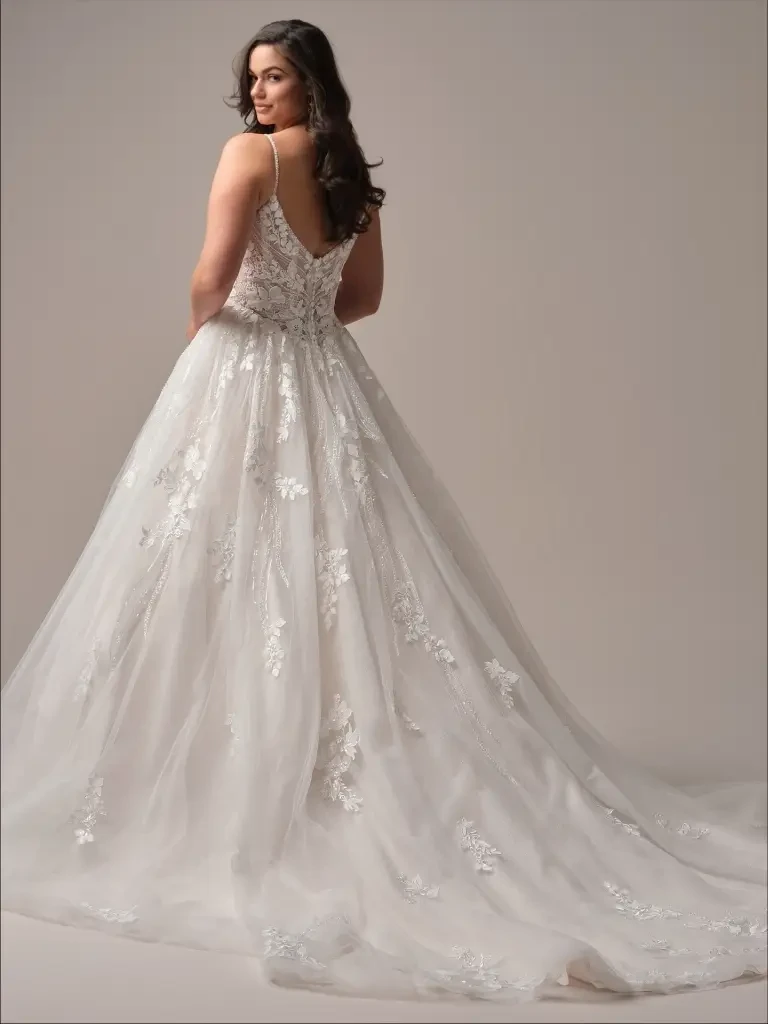 A woman in a white wedding dress with delicate lace details, featuring a fitted bodice and a flowing skirt, standing gracefully against a neutral background.