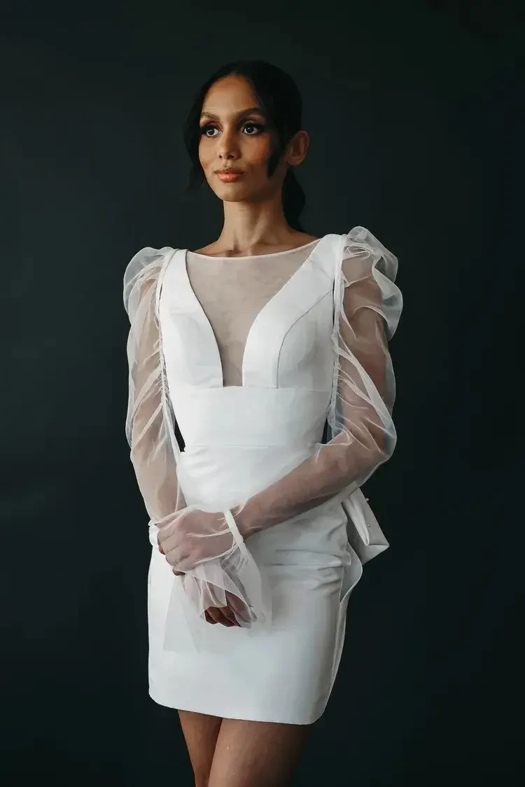 A woman in an elegant white dress with sheer puff sleeves poses against a dark background. Her expression is calm and confident, exuding a sophisticated tone.