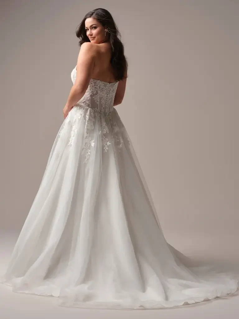 A woman wearing a strapless wedding dress with a fitted bodice and a flowing tulle skirt, standing elegantly against a neutral background.
