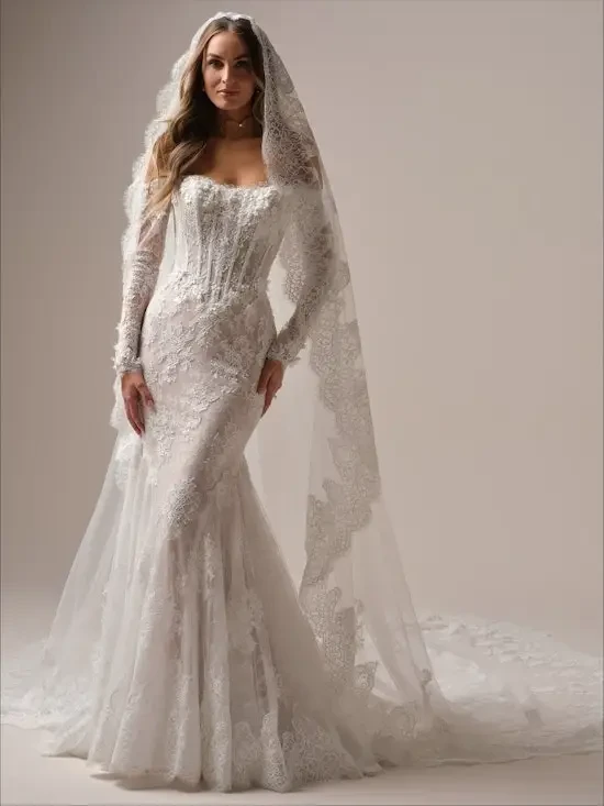A woman wearing an elegant wedding dress with intricate lace detailing and a long veil, standing gracefully.