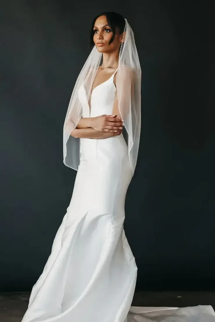 A bride wearing a sleek white wedding gown and a long veil, standing elegantly against a dark background.